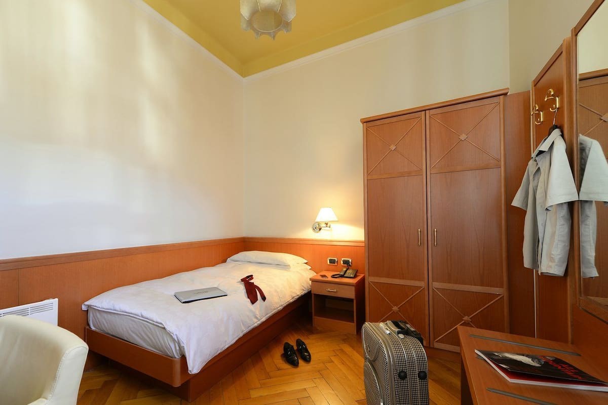 A hotel room with a single bed, a desk with a chair, a wardrobe, and a lamp.