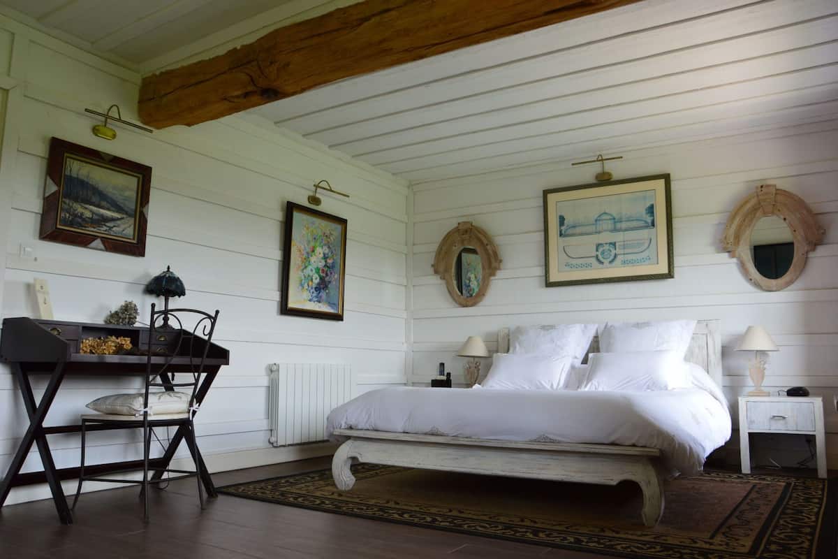 A bedroom with a bed, a desk, and a chair. There are paintings on the walls and a mirror above the bed.