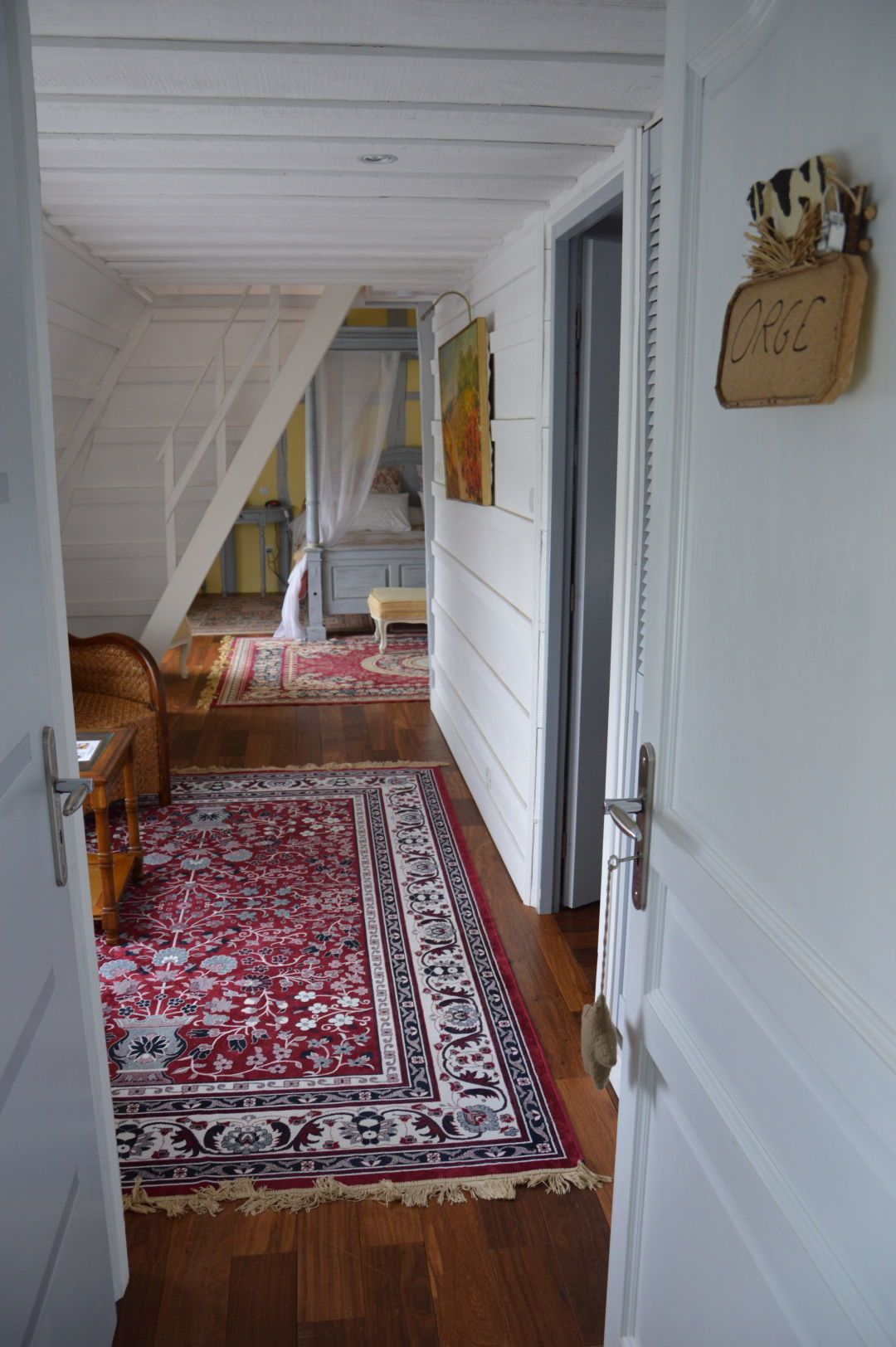 A hallway with a red and white patterned rug, a wooden bench, and a bed with a canopy in the background.