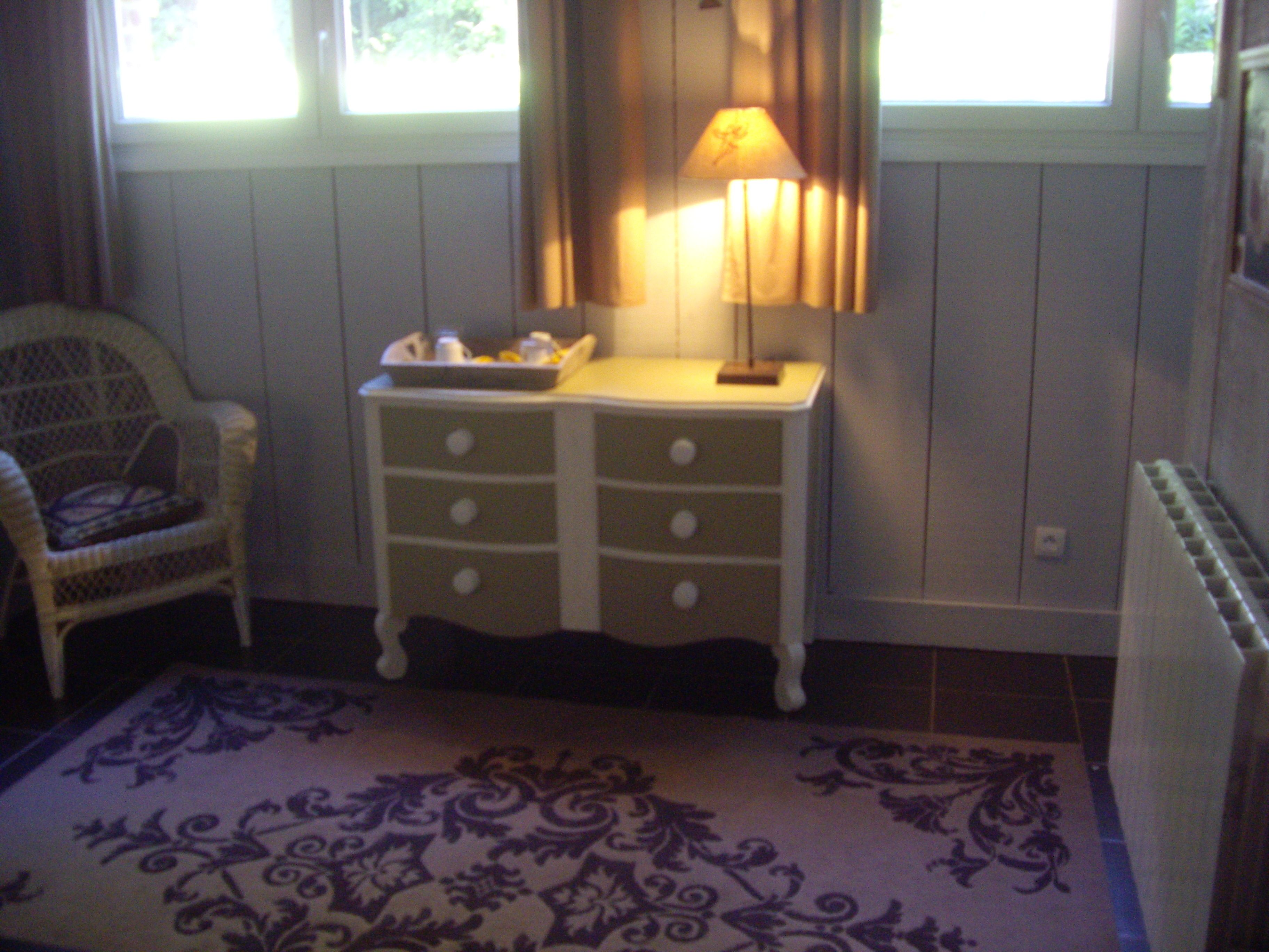 Quarto com cômoda branca, cadeira de vime, tapete estampado e janela com cortinas.