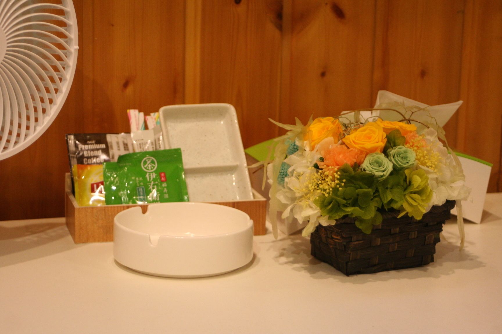A table with a white ceramic bowl, a basket of flowers, and a fan.