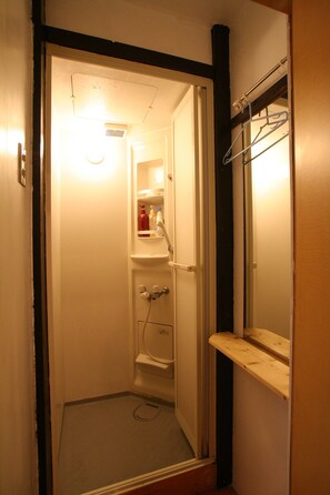 A small, narrow bathroom with a shower, a mirror, and a shelf with toiletries.