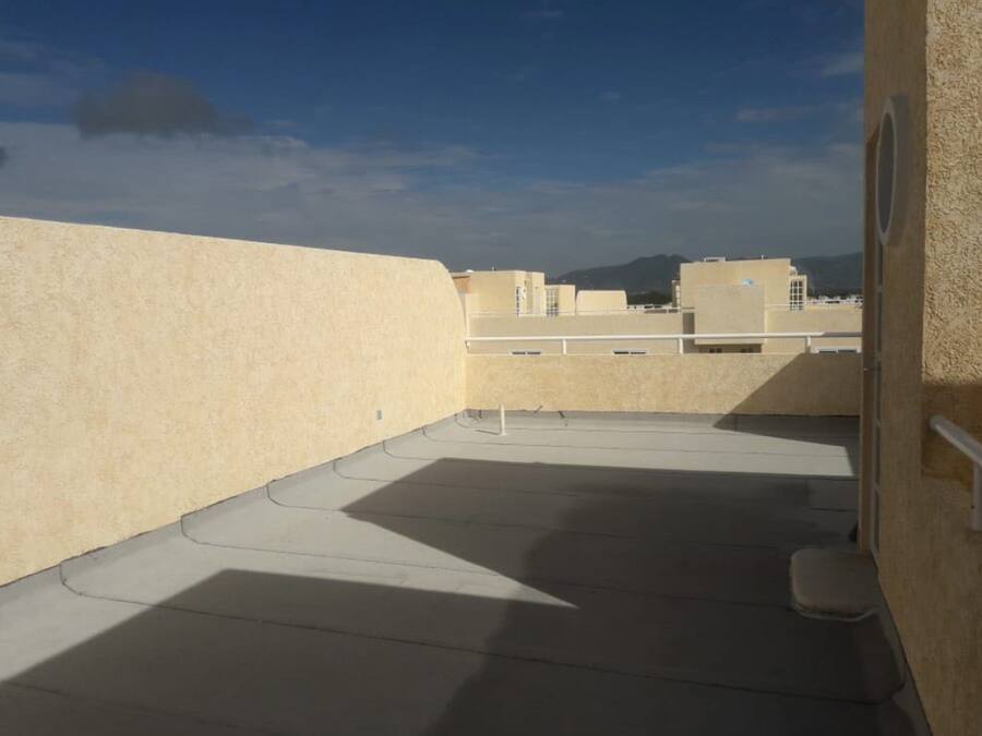 A rooftop terrace with a concrete floor, beige walls, and a view of other buildings and mountains in the distance.