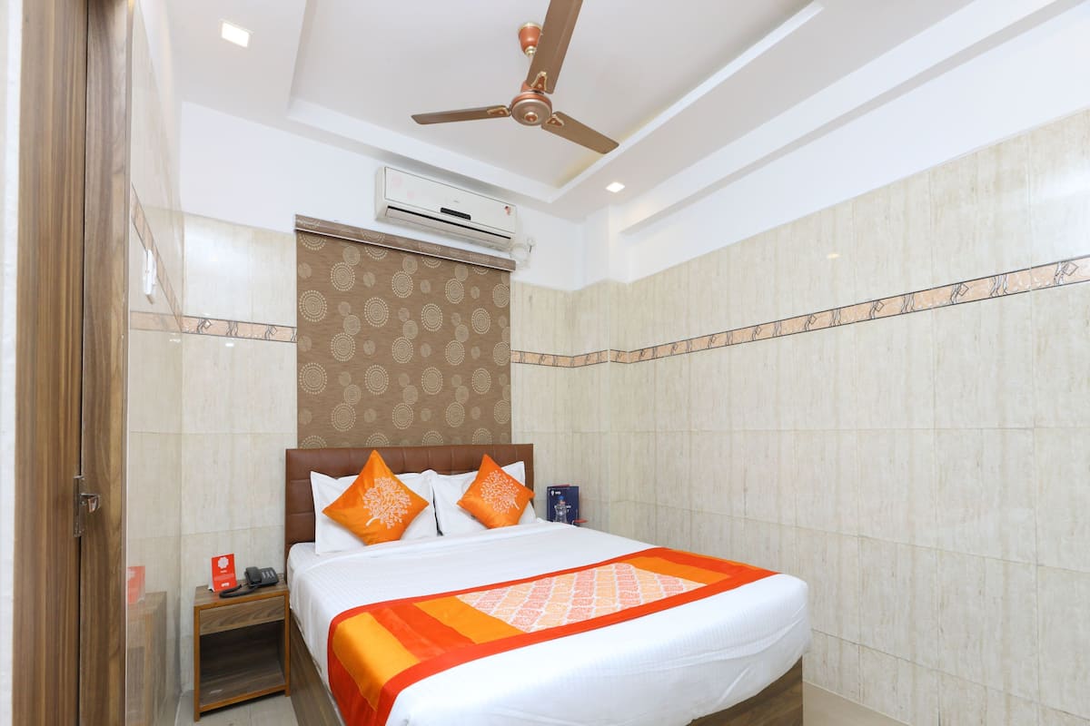 Una habitación de hotel con una cama, un equipo de aire acondicionado, un ventilador de techo y una mesita de noche con un teléfono y un cartel rojo.
