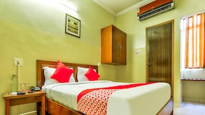 Habitación de hotel con cama de madera, ropa de cama blanca y roja, mesita de noche con teléfono y botellas de vidrio, armario de madera y puerta con ventana.