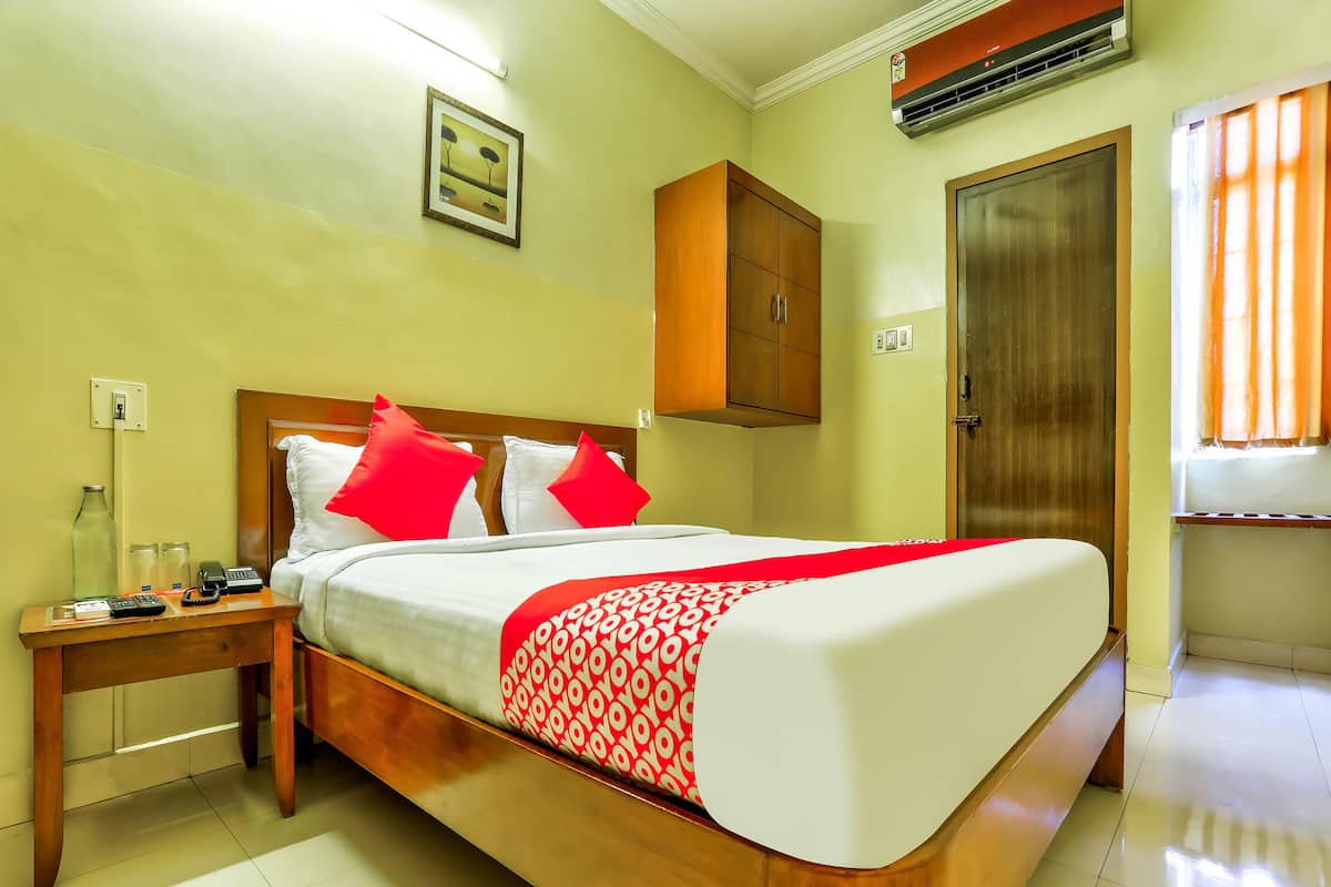 Habitación de hotel con cama de madera, ropa de cama blanca y roja, mesita de noche con teléfono y botellas de vidrio, armario de madera y puerta con ventana.