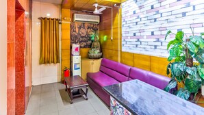 Un área de recepción con un sofá morado, una pequeña mesa de madera, una estatua decorativa de Buda y una pared con diseño de ladrillo.