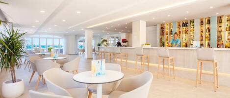 A modern bar area with white chairs and tables, a well-lit ceiling, and a bar stocked with various bottles.