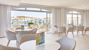 A spacious indoor area with multiple round tables and chairs, large windows offering a view of the beach and umbrellas outside.