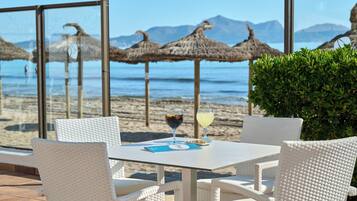 A table with white wicker chairs, a glass of red wine, and a menu on a terrace overlooking a beach with thatched umbrellas and mountains in the distance.