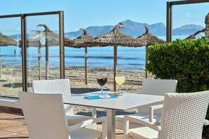 A table with white wicker chairs, a glass of red wine, and a menu on a terrace overlooking a beach with thatched umbrellas and mountains in the distance. - Grupotel Dunamar (Santa Margalida)