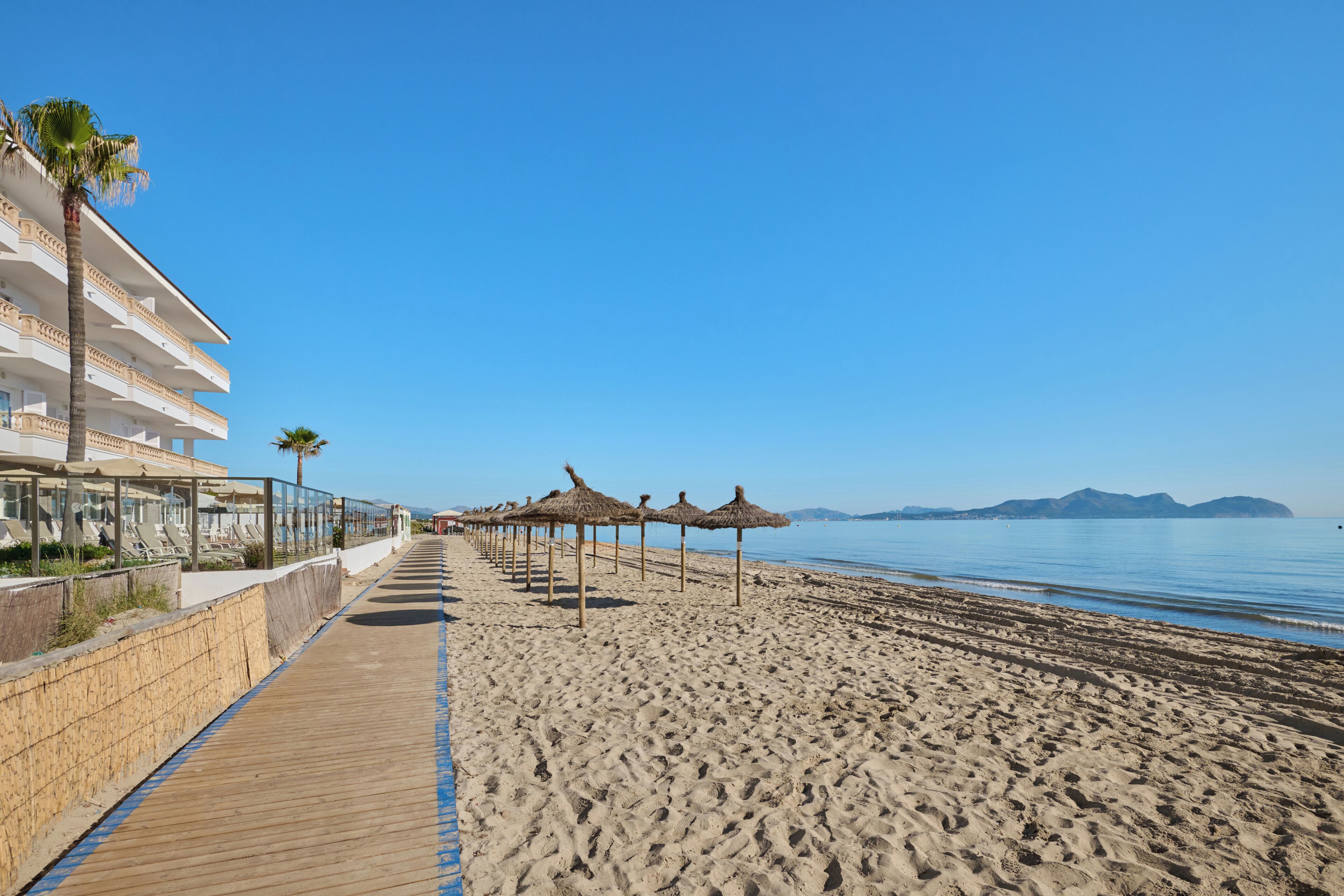 Een hotel aan het strand met een houten pier, strandparasols en een helder uitzicht op zee.