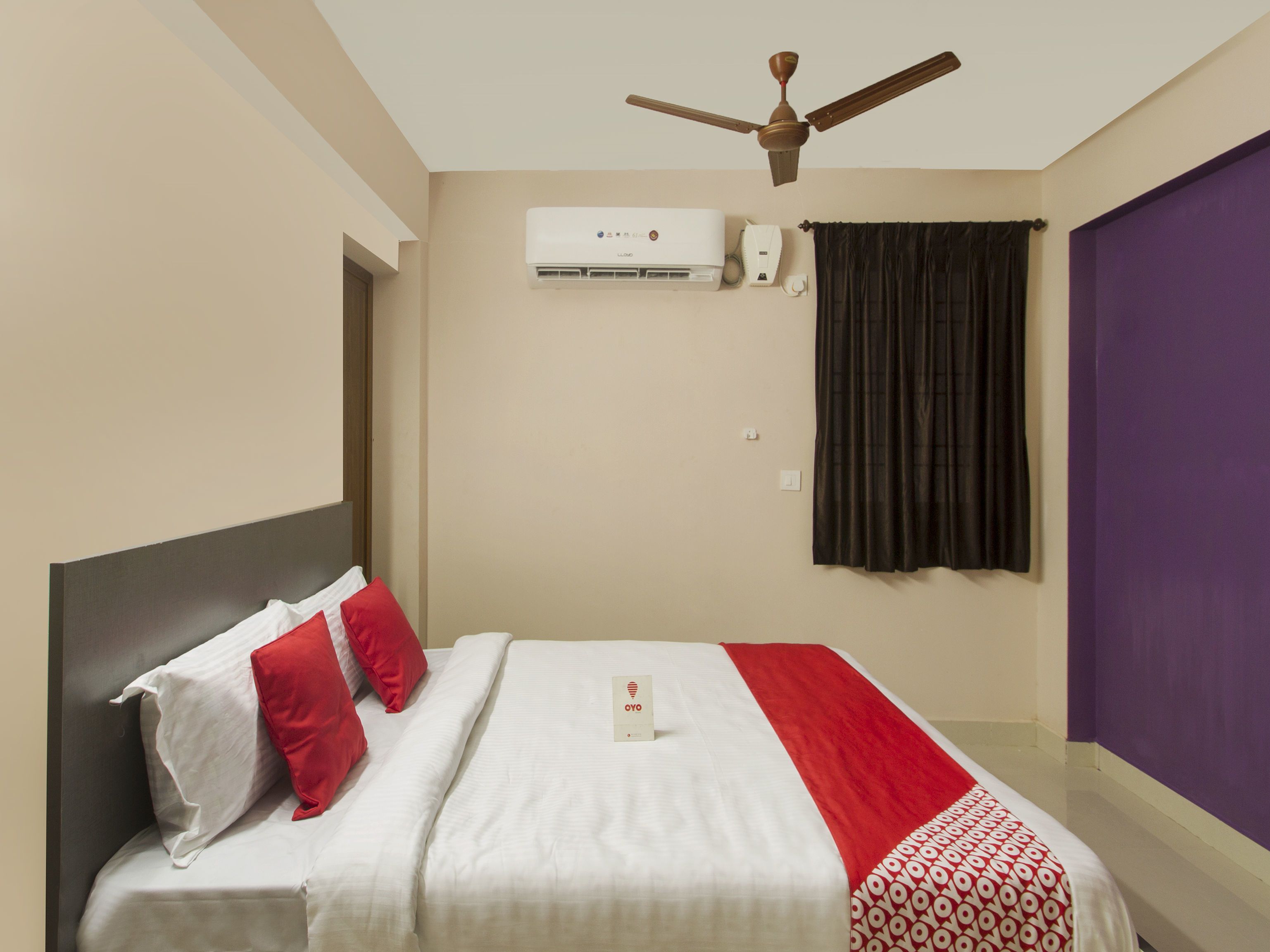 A hotel room with a bed, a ceiling fan, an air conditioning unit, and a window with curtains.