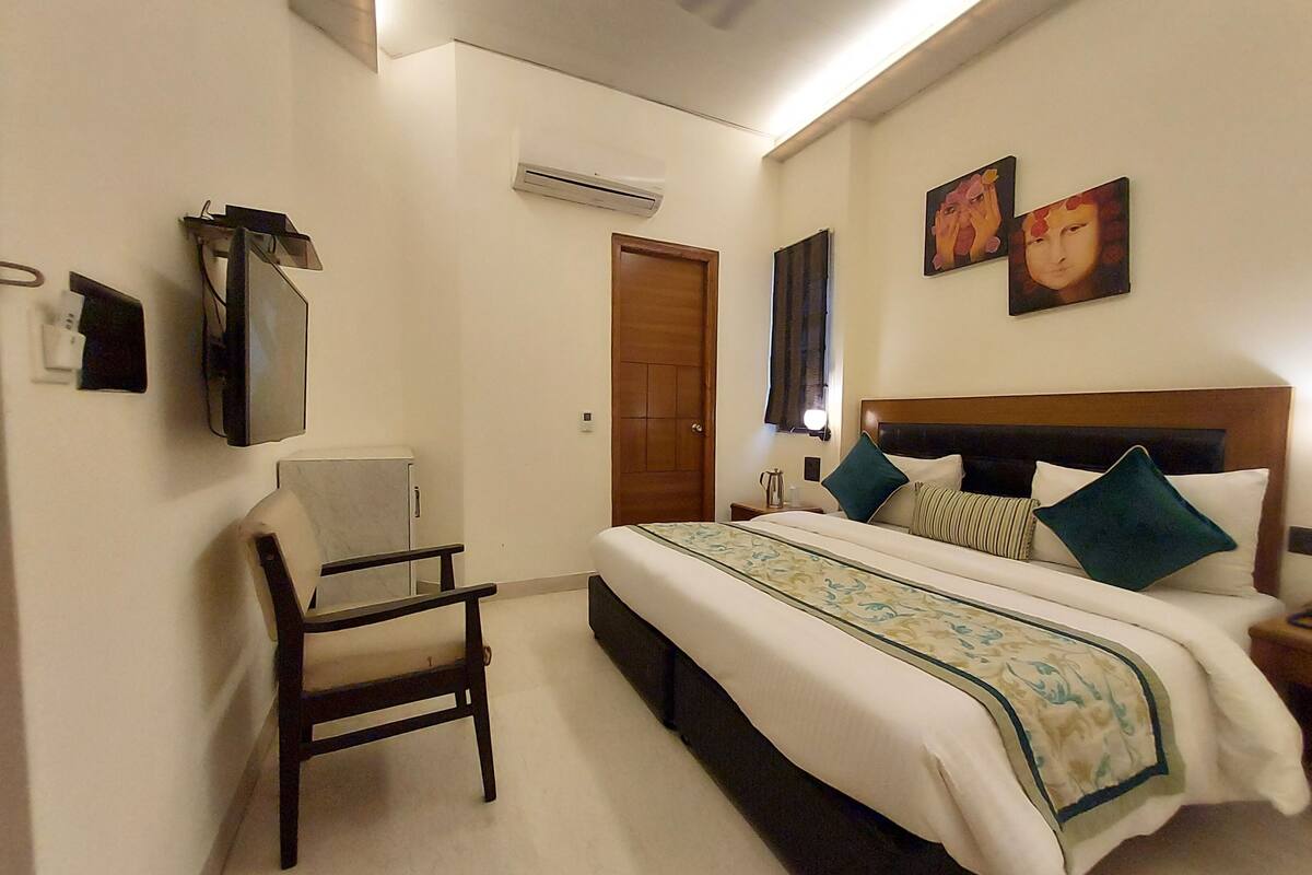 A hotel room with a bed, a chair, a desk, an air conditioning unit, and artwork on the walls.