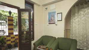 Una zona de recepción con una puerta de madera, dos sillones verdes con cojines estampados, un cartel con una estatua de Buda y un suelo de baldosas con plantas en macetas.