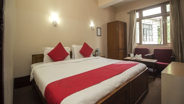 Habitación de hotel con una cama grande, dos almohadas rojas, un armario de madera, un escritorio con una silla y una ventana con cortinas.