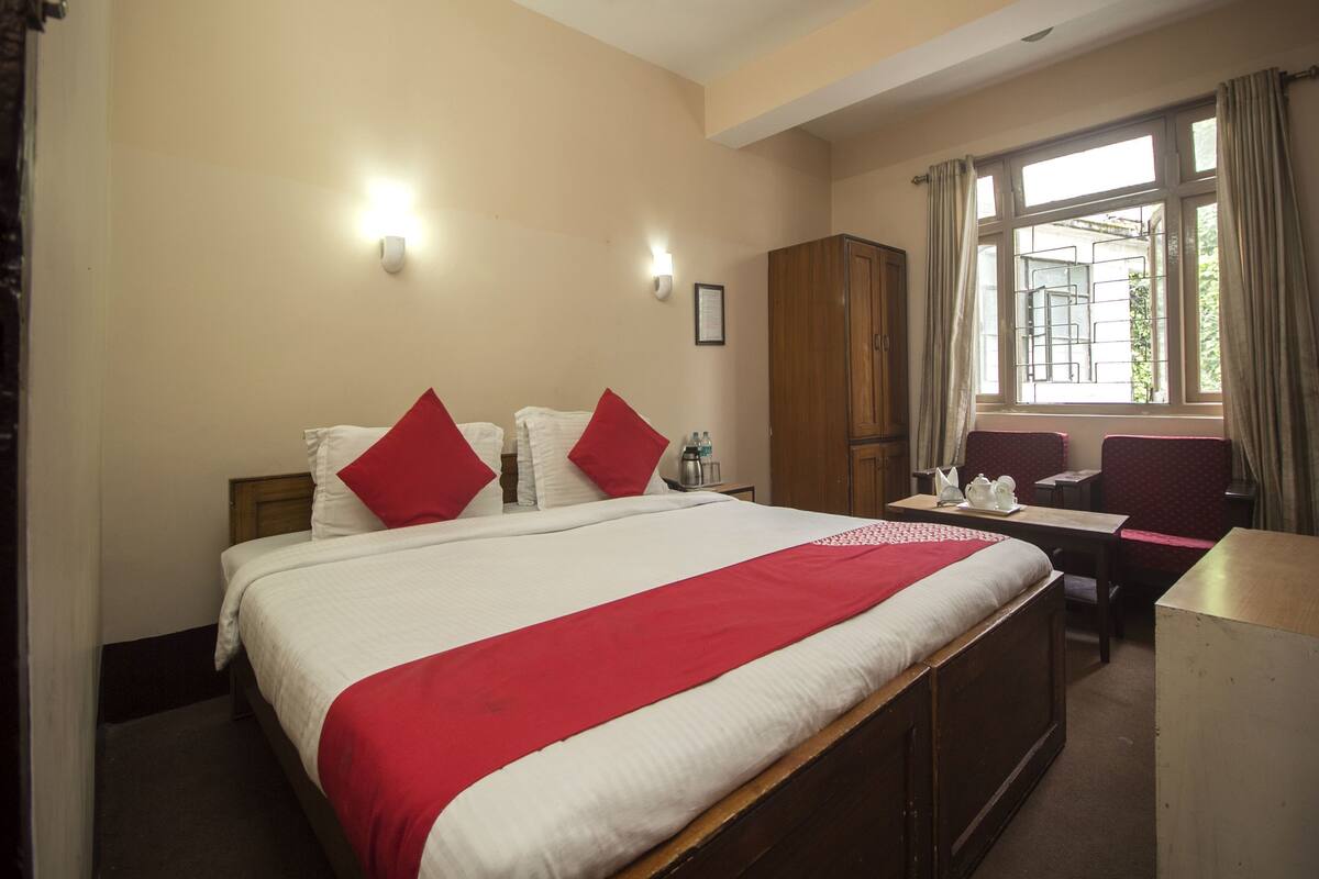 Habitación de hotel con una cama grande, dos almohadas rojas, un armario de madera, un escritorio con una silla y una ventana con cortinas.