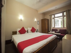 Habitació d'hotel amb un llit gran, dos coixinets vermells, un armari de fusta, un escriptori amb una cadira i una finestra amb cortines.