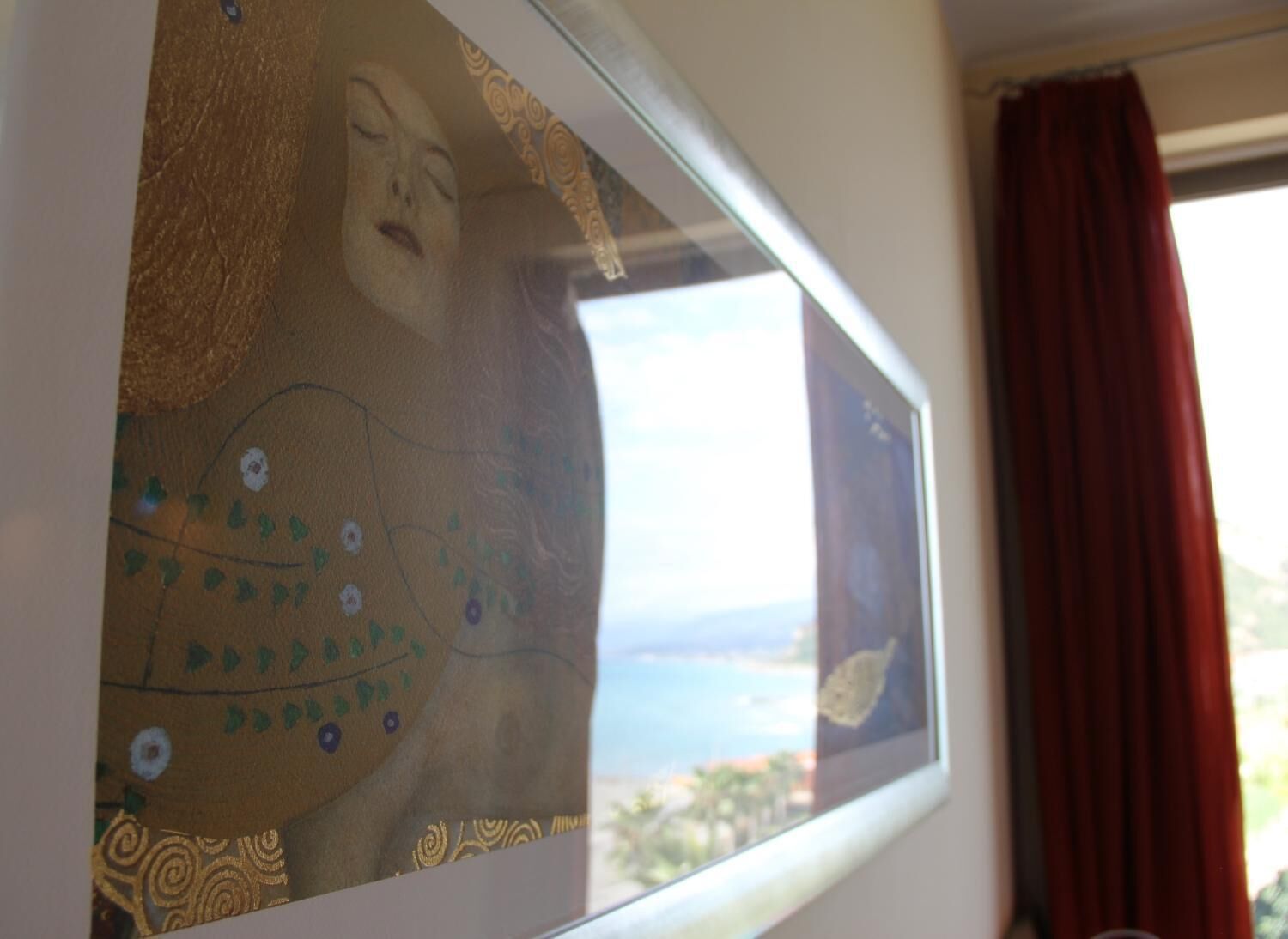 Ein Fenster mit Blick auf eine Küstenlandschaft, ein roter Vorhang und ein gerahmtes Kunstwerk mit floralen Mustern.