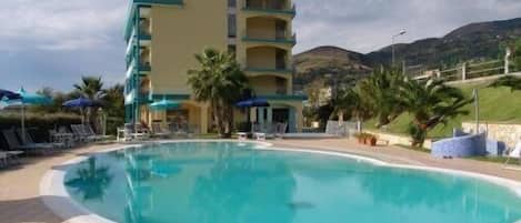 A hotel with a pool, multiple floors, and a mountainous backdrop.