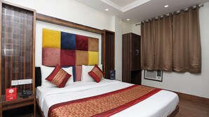Hotelkamer met groot bed, houten hoofdbord, kleurrijk wandpaneel, bruin gordijn en klein nachtkastje met telefoon en rood bordje.