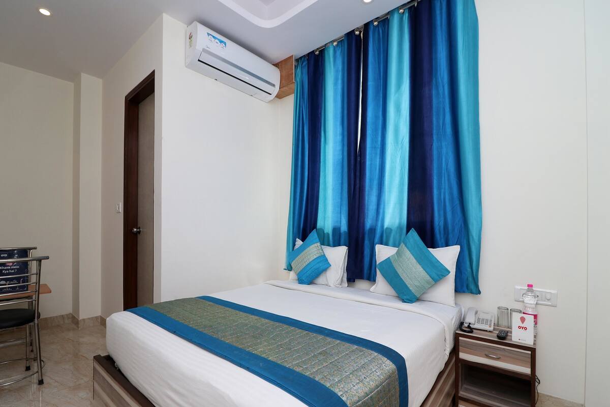 A hotel room with a bed, a nightstand, a desk, an air conditioning unit, and a curtain.