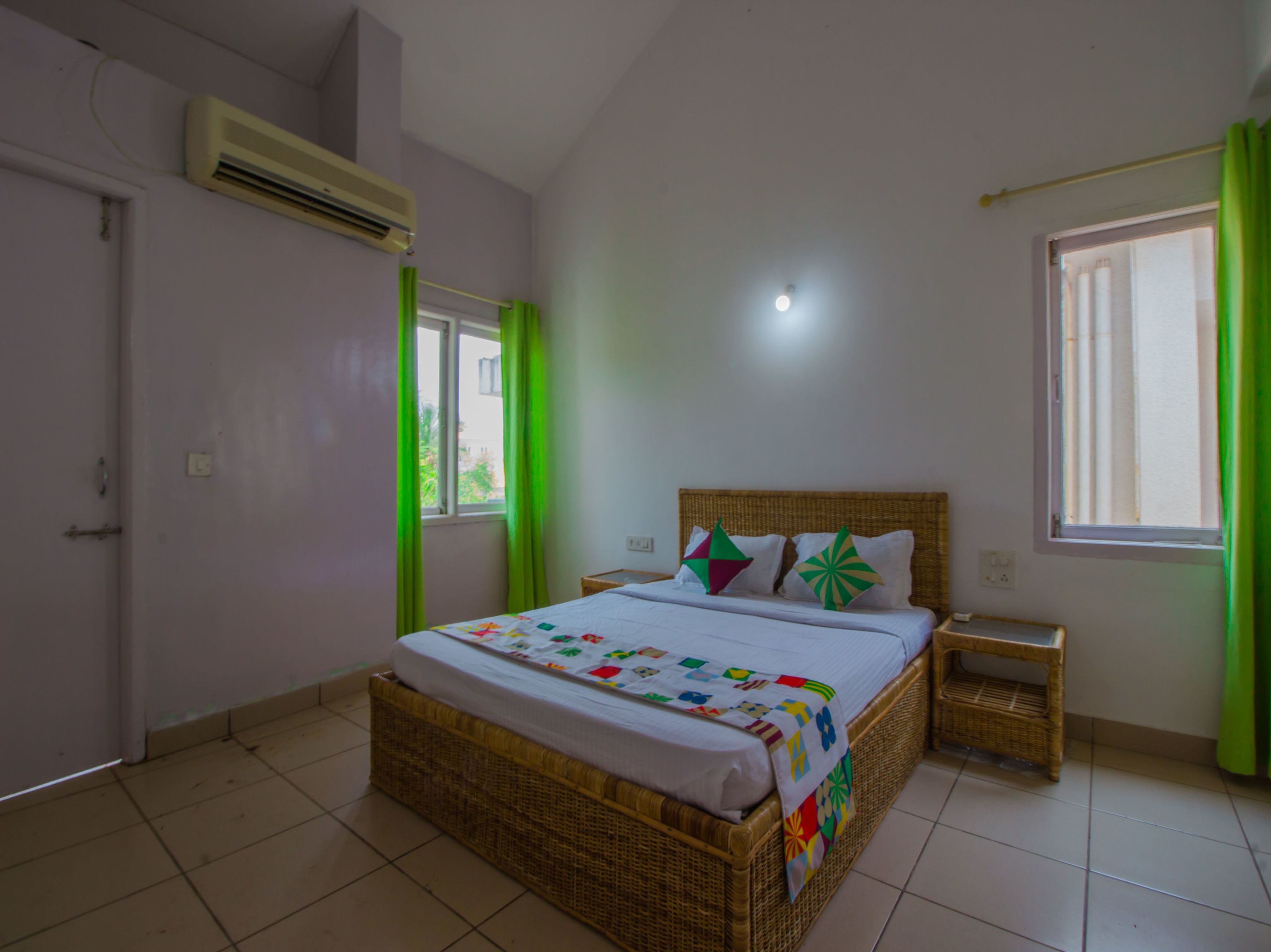 A bedroom with a wicker bed, a nightstand, a window with green curtains, and an air conditioning unit on the wall.