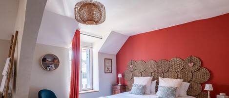 A bedroom with a red accent wall, a large bed with multiple pillows, a blue chair, a wooden bench, and a unique woven headboard.