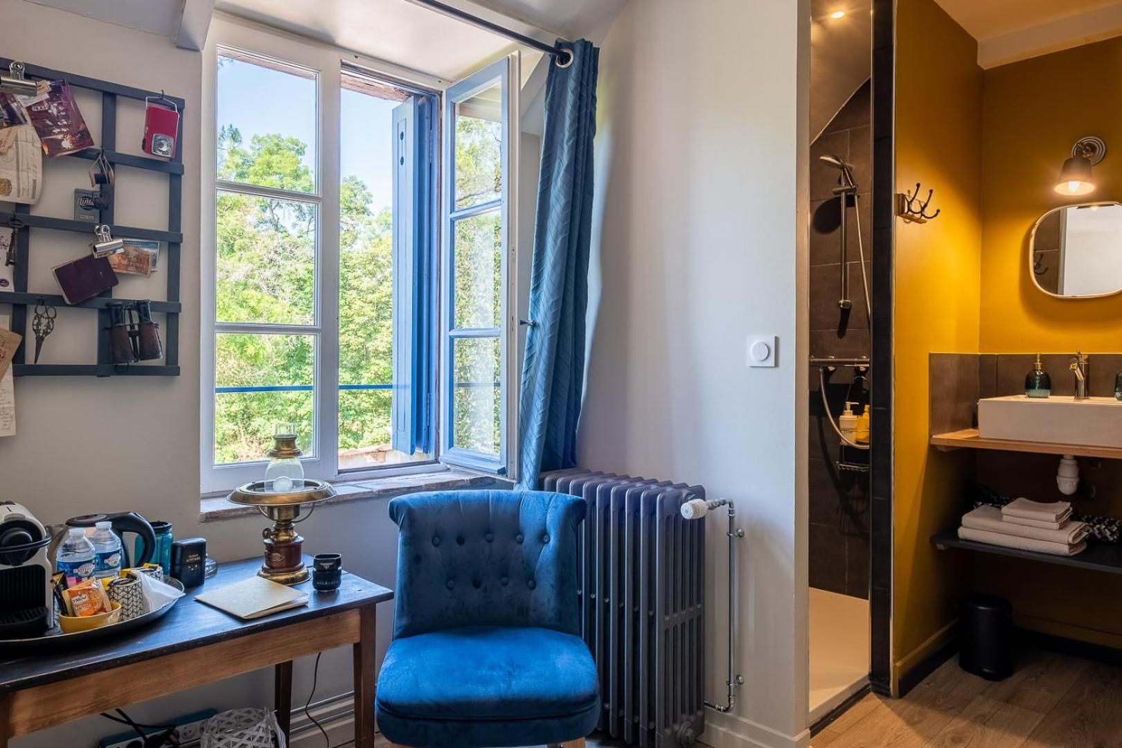 Quarto com cadeira azul, radiador, prateleira com artigos de higiene e janela com cortinas.