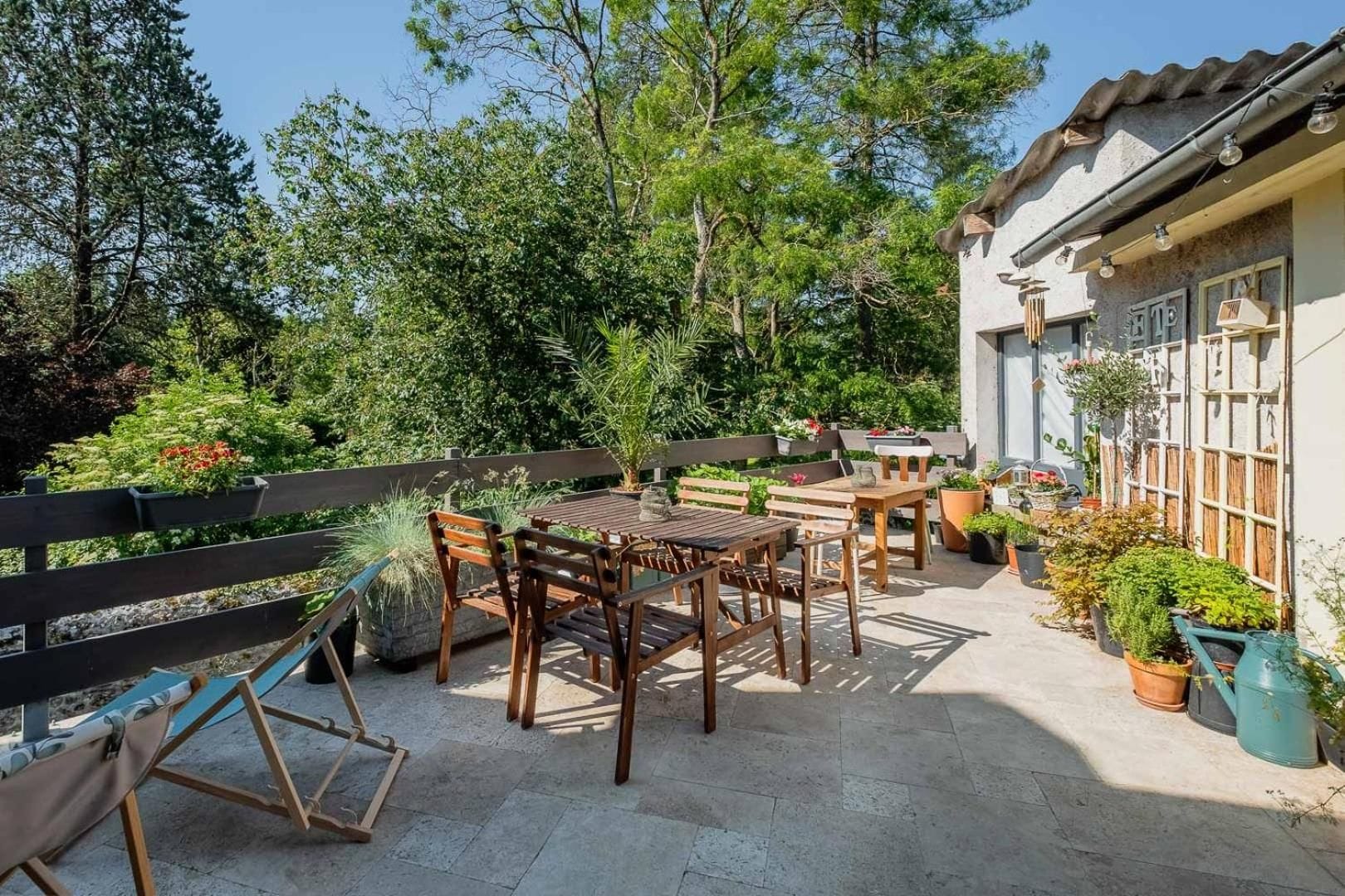 An outdoor patio area with a wooden table and chairs, surrounded by potted plants and a wooden fence.