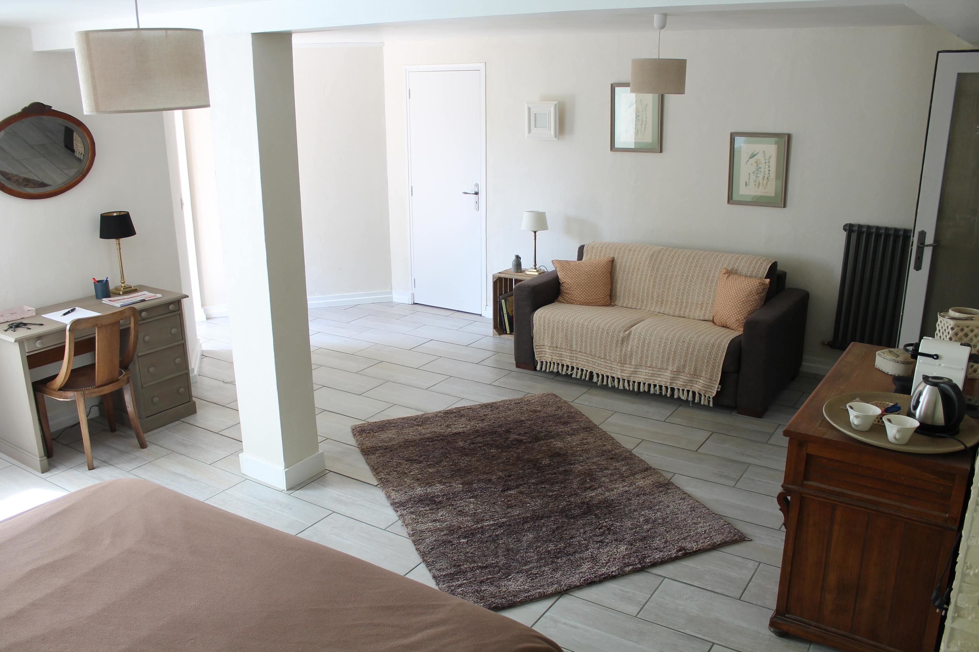 Une pièce spacieuse comprenant un lit, un bureau avec une chaise, un canapé et une table basse. Une porte donne accès à une autre pièce et un radiateur est fixé au mur.