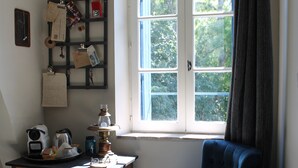 A room with a window, a chair, a table with various items, and a shelf with knick-knacks. - A La Lettre Thé (Valencisse)