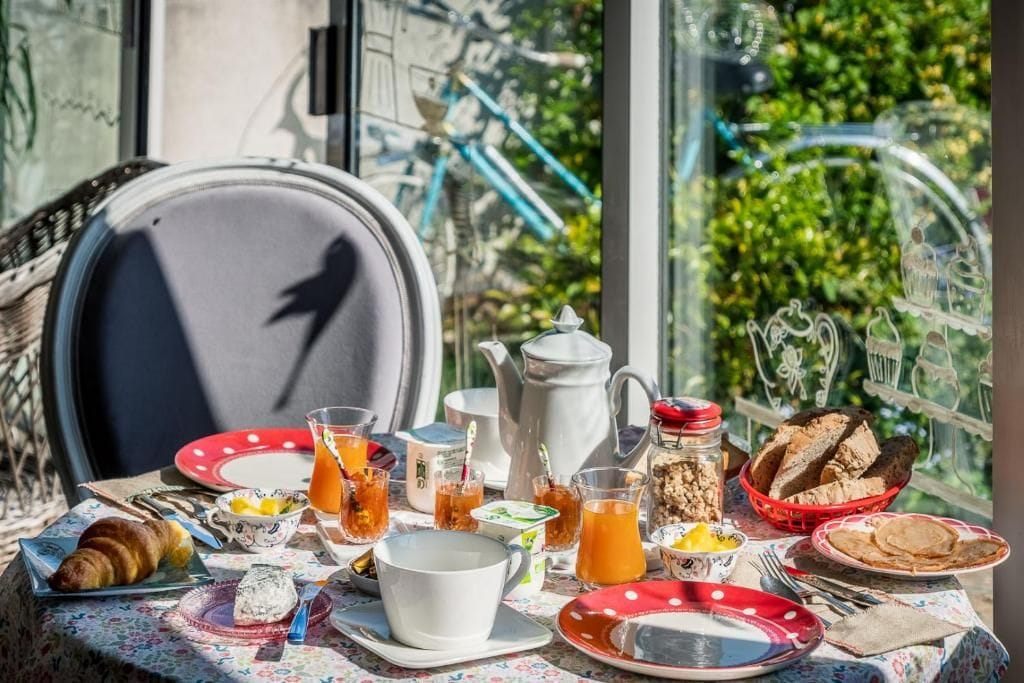 Une table dressée pour le petit-déjeuner, avec un croissant, du thé et du pain, et en toile de fond, une vue sur la verdure et des vélos à l'extérieur.