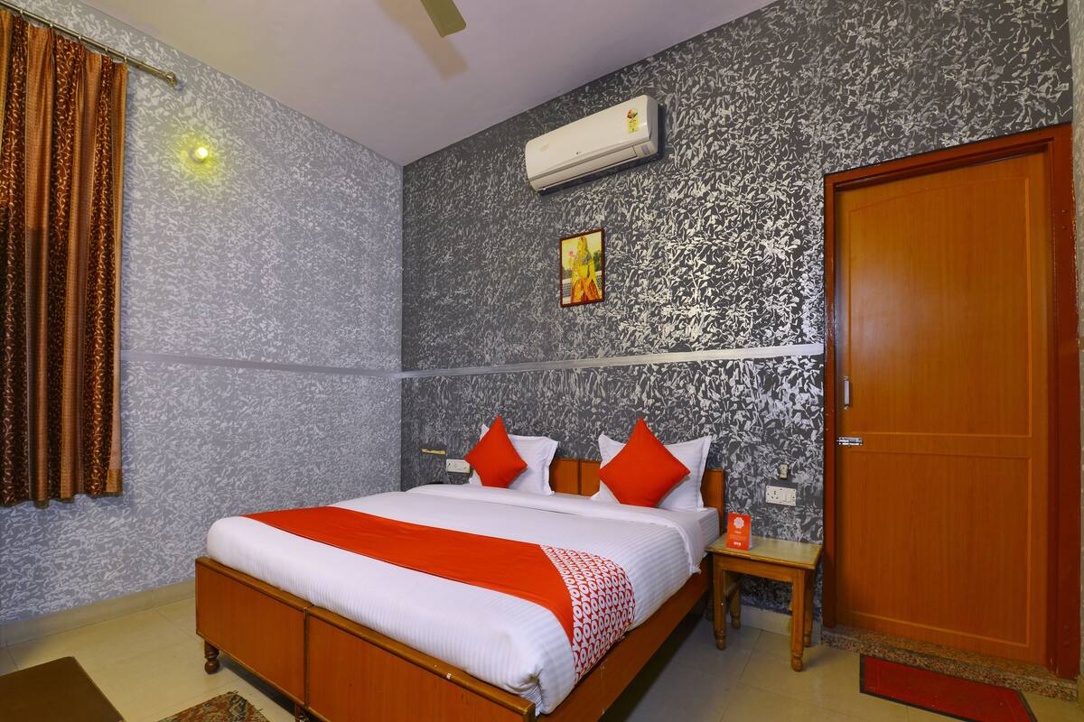 A bedroom with a wooden bed, red and white bedding, a patterned wall, an air conditioner, a wooden door, and a small bedside table.