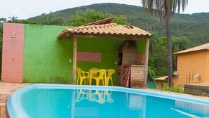 Una piscina con borde de baldosas, un área de estar techada con sillas amarillas, y una estructura de ladrillo con techo de tejas.