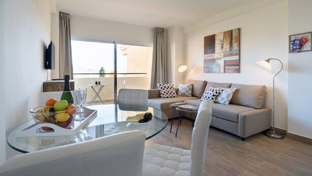 Een moderne woonkamer met een glazen eettafel, een zithoek en een groot raam met gordijnen.