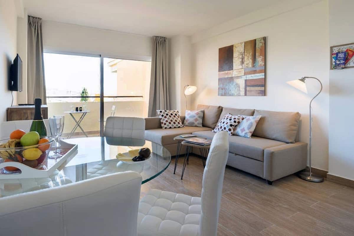 A modern living room with a glass dining table, a sofa set, and a large window with curtains.