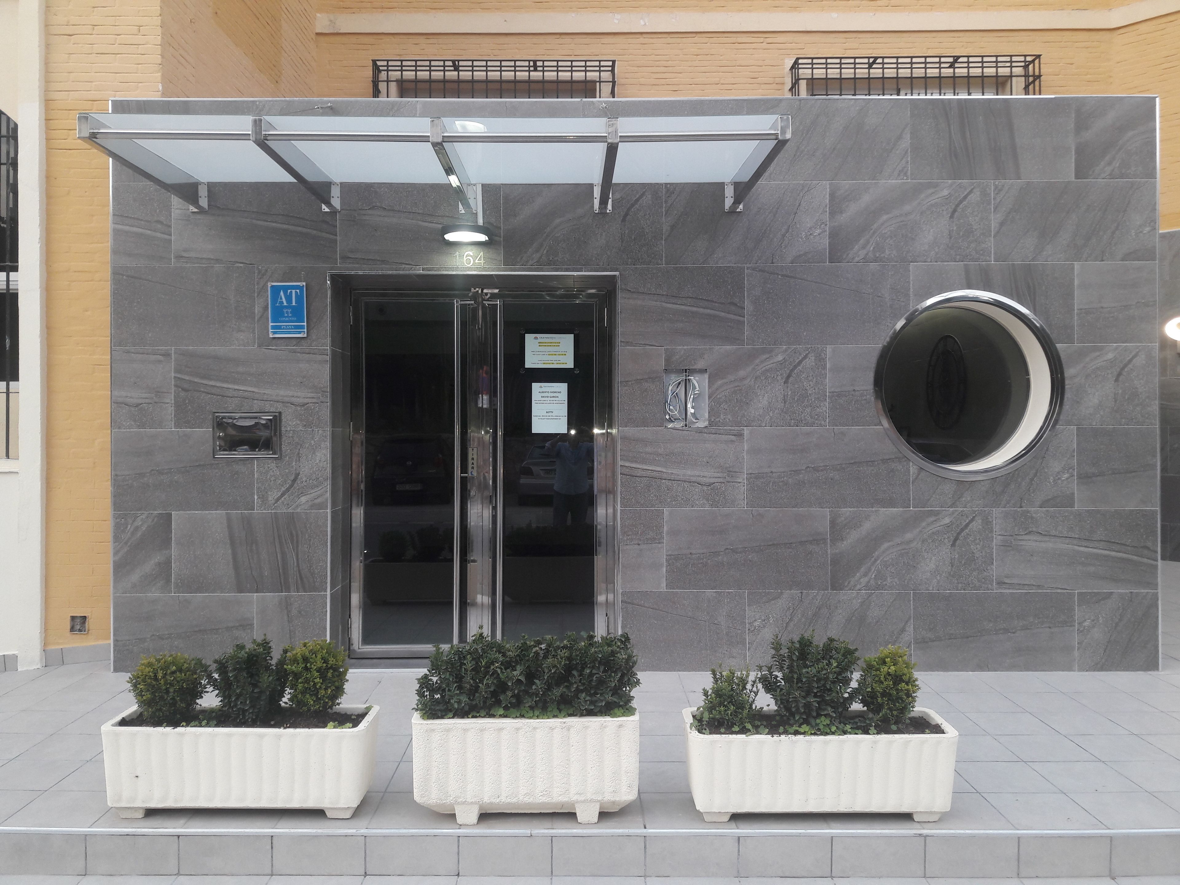 A building entrance with a glass door, a number '164' sign, a round mirror, and three white planters with shrubs.