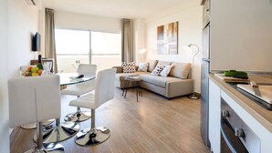 Een moderne woonkamer met een bank, een glazen eettafel en een keukenhoek met een koelkast en een snijplank.