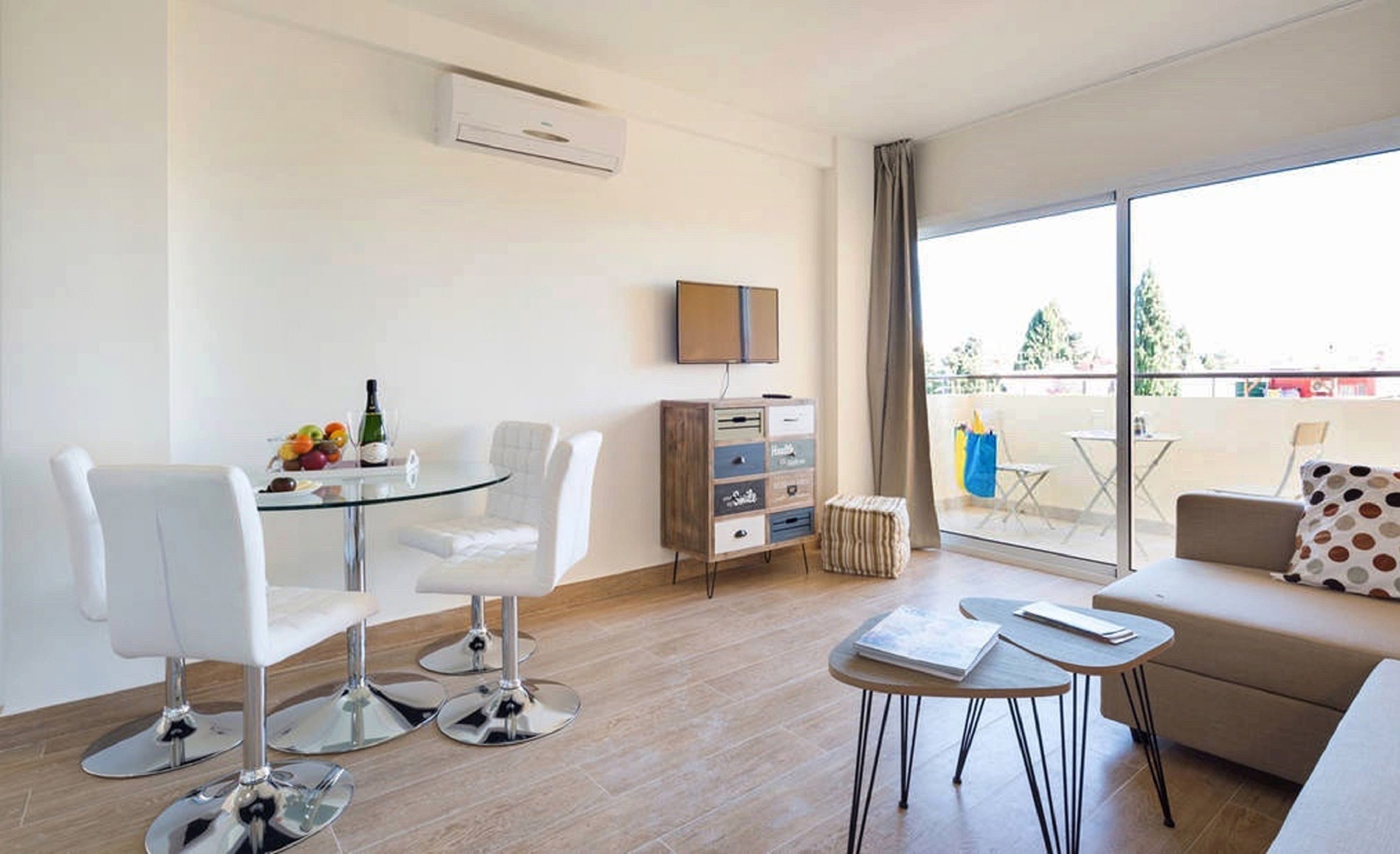 A modern living room with a glass table, white chairs, a sofa, a TV, and a balcony with outdoor furniture.