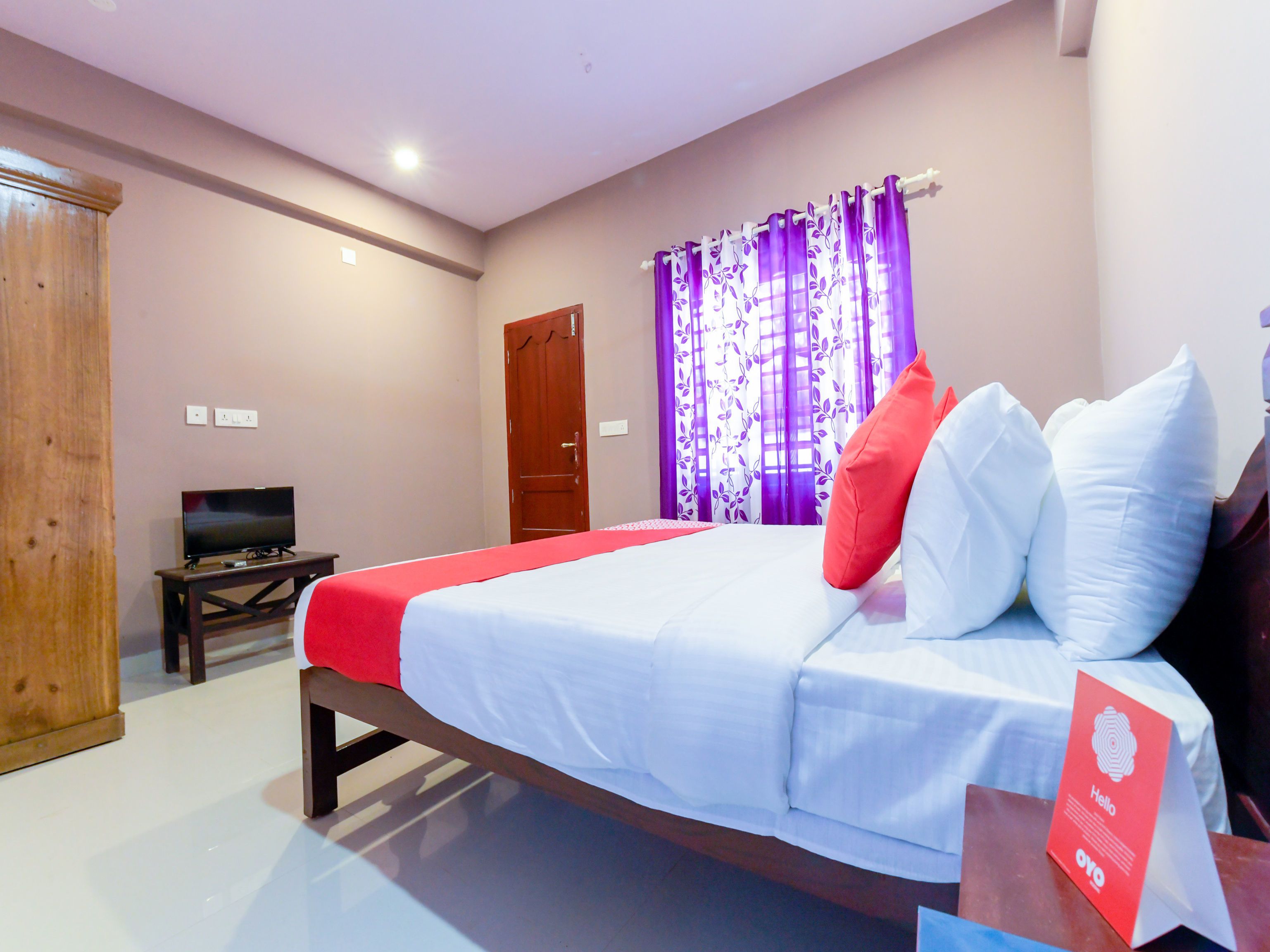 A hotel room with a bed, a TV, a wooden wardrobe, and purple curtains.