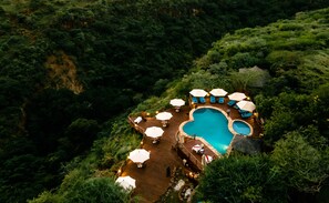 Luchtbeeld van een resort met een zwembad, omringd door ligbedden en parasols, met op de achtergrond een dicht bos.