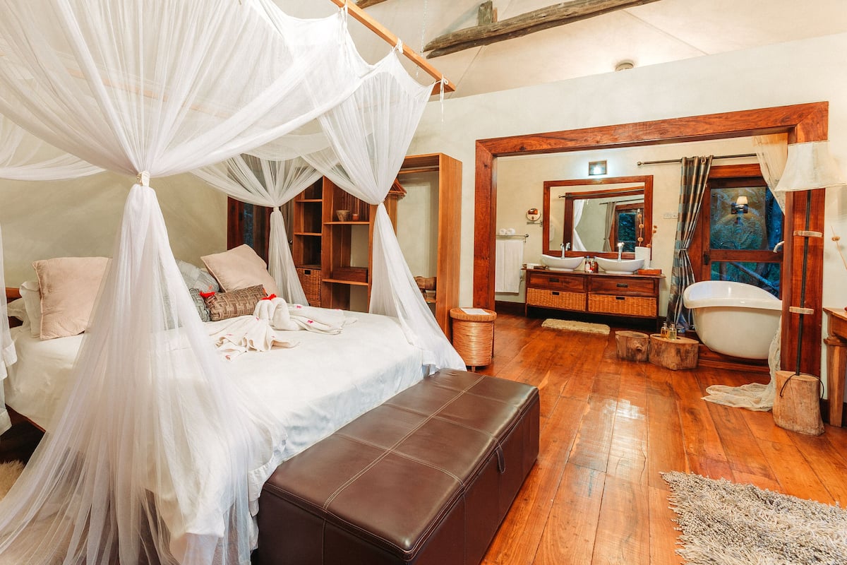 Een slaapkamer met een hemelbed, houten vloeren, een grote spiegel en een houten bankje.