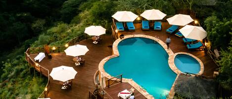 An outdoor swimming pool surrounded by lounge chairs and umbrellas, set on a wooden deck with a forest backdrop.