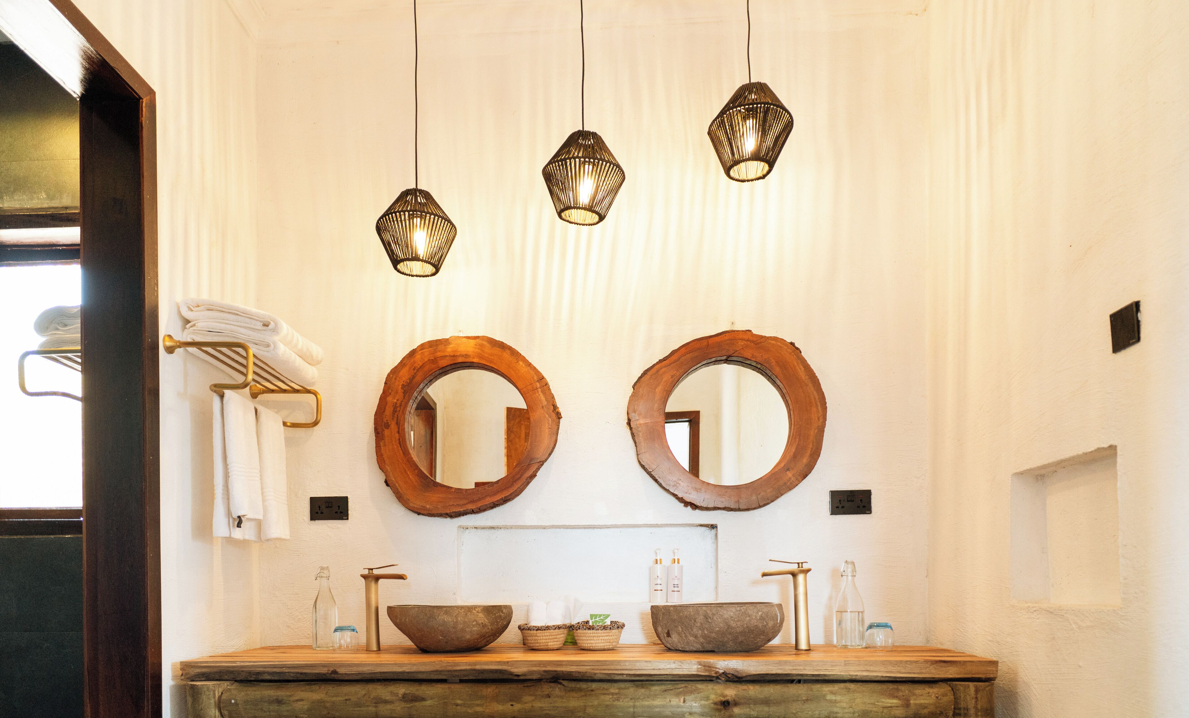 A bathroom with two wooden-framed mirrors, a wooden vanity with a bowl sink, a towel rack with folded towels, and three hanging pendant lights.