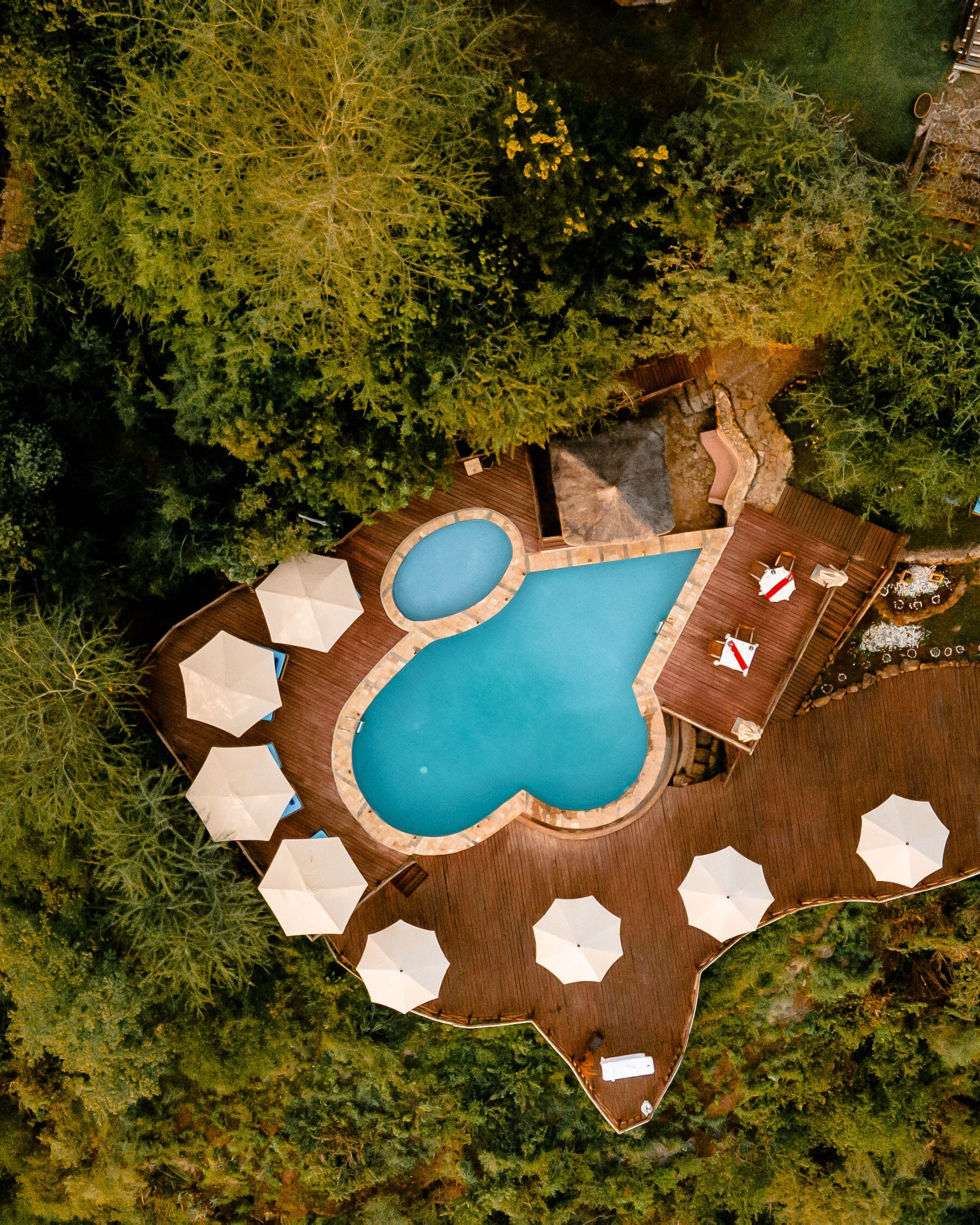 An aerial view of a resort with a central swimming pool surrounded by wooden deck areas and white umbrellas.