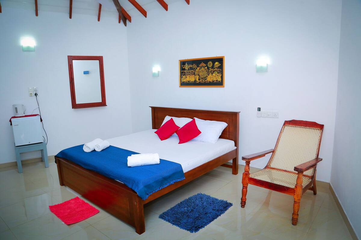 A neatly made bed with a blue and red blanket, a wooden chair, and a small table with a kettle.