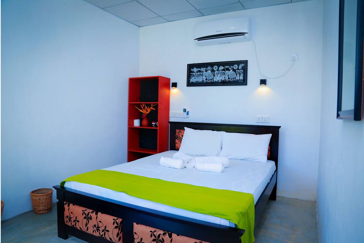 A neatly made bed with white linens and a green throw, a red bookshelf, and a wall-mounted air conditioner.