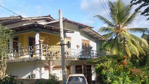 Una casa a due piani con un balcone, un'auto parcheggiata e una palma sullo sfondo.
