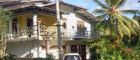 Una casa de dos pisos con un balcón, un coche aparcado y una palmera al fondo.
